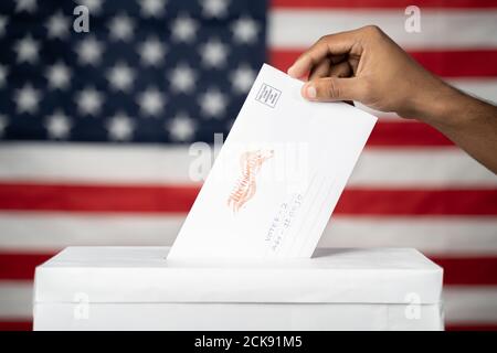Maski, Inde 14 septembre, 2020 : concept de Mail en vote aux élections américaines - mains déposant le courrier à l'intérieur de l'urne avec nous drapeau comme arrière-plan. Banque D'Images