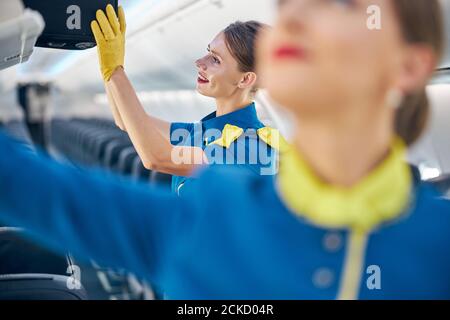 Portrait en gros plan d'une femme souriante et gaie portant un préposé à l'avion uniforme bleu tenant à la main sac à roulettes noir pendant la mise il sur la cabine com Banque D'Images