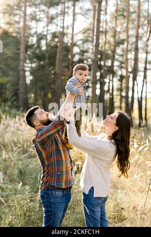 Portrait de famille en plein air de jeunes parents heureux, portant des vêtements décontractés élégants, s'amuser et soulever leur petit fils mignon, pendant l'entrée Banque D'Images