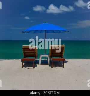 Vue arrière d'une paire de chaises longues en bois lattes sur une plage tropicale de sable blanc, sous un parasol bleu. Banque D'Images