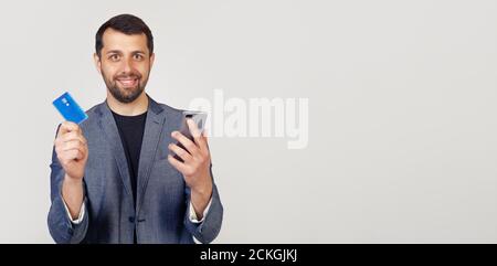 Jeune homme d'affaires avec une barbe dans une veste, beau homme utilisant une carte de crédit pour payer en ligne à l'aide d'un smartphone. Portrait d'un homme sur fond gris. Banque D'Images