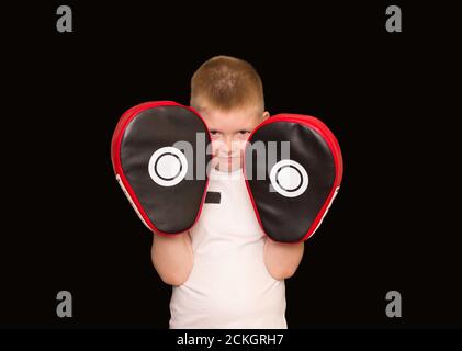 Boxe pour enfants, sports. Un garçon dans un t-shirt blanc avec des pattes de boxe rouges sur fond noir. Banque D'Images