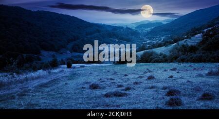 route de campagne à travers le champ rural la nuit. paysage d'été suburbain dans les montagnes en pleine lune lumière. village dans la vallée éloignée. jour nuageux Banque D'Images