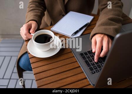 Femme en laine manteau travaillant dans un café de rue avec ordinateur portable. Une tasse de café sur une table en bois. Banque D'Images