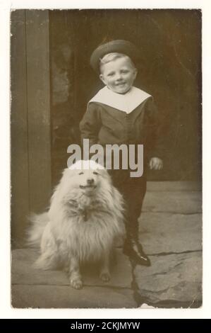 Au début des années 1900, carte postale d'un adorable petit garçon édouardien portant un costume de marin avec son chien de compagnie, peut-être la race de Samoyed, debout sur des dalles, à l'extérieur de sa maison, vers 1910, Royaume-Uni Banque D'Images