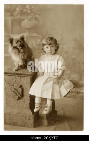 Au début des années 1900, la carte postale du portrait d'un jeune enfant (fille ou garçon qui s'habit de la même façon dans ces temps) avec chien d'animal de compagnie, vers 1910, R. Wilson, Carlton Studio, Consett, Comté de Durham, Angleterre, Royaume-Uni Banque D'Images