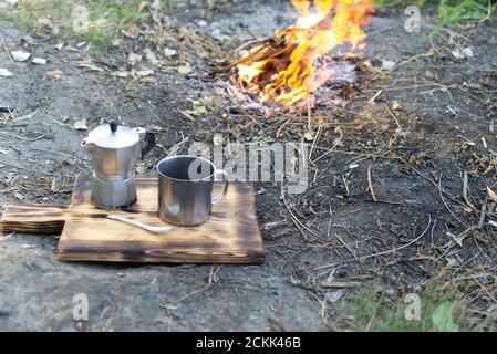Café chaud dans une tasse de métal avec une machine à café geyser sur une planche à bois près du feu. Week-end d'automne. Mise au point sélective. Orientation horizontale. Banque D'Images