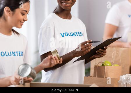 Concentration sélective des volontaires afro-américains tenant le stylo et le presse-papiers près d'une femme asiatique avec des boîtes de conserve et des paquets dans la charité centre Banque D'Images