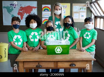 Portrait d'une enseignante et d'un groupe d'enfants portant le visage masques avec récipient de recyclage à l'école Banque D'Images