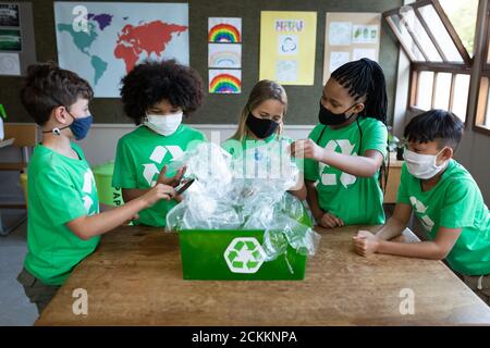 Portrait d'un groupe d'enfants portant un masque facial touchant des plastiques article dans le conteneur de recyclage de la classe Banque D'Images