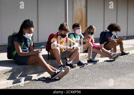 Groupe d'enfants portant un masque facial avec un smartphone lorsqu'ils sont assis ensemble Banque D'Images