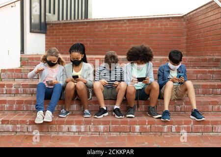 Groupe d'enfants portant un masque facial avec un smartphone lorsqu'ils sont assis sur les escaliers ensemble Banque D'Images