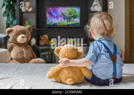 Un joli petit garçon et son ours en peluche regardant la télé s'asseoir sur un canapé dans le salon à la maison Banque D'Images