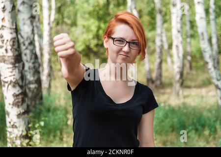 Fille de gingembre montrer le pouce vers le haut. Concept de désapprobation sociale. Jeune belle femme en t-shirt noir et lunettes dans le parc. Banque D'Images