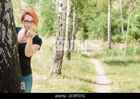 Concept d'observation et de surveillance avec espace de copie. Belle fille de gingembre se tenir derrière l'arbre et de pointer le doigt à l'appareil photo. Jeune femme en bouleau d'été pour Banque D'Images