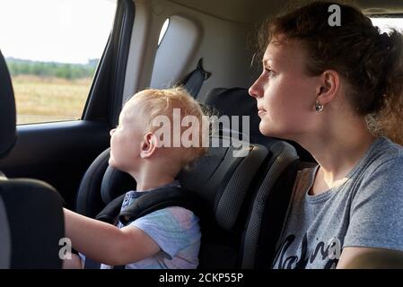 Un petit garçon est assis dans un siège de voiture près de sa mère. Maman et bébé regardent la fenêtre. Sécurité enfant dans la voiture. Banque D'Images