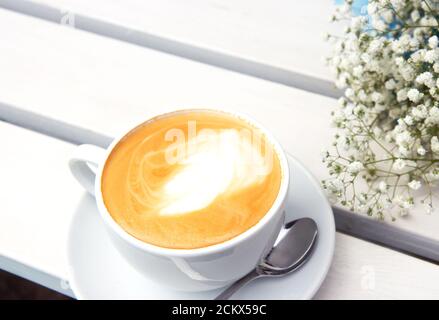 Cappuccino dans une tasse en céramique blanche et bouquet de petites fleurs blanches sur un banc en bois. Café rustique en plein air le matin. Routine du matin. Gypsophila Banque D'Images