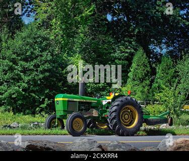 Un tracteur John Deere classique avec une plate-forme de tonte fixée à l'arrière est stationné sur le côté de la route. En arrière-plan se trouvent des arbres verts luxuriants Banque D'Images