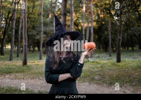 Une jeune femme en robe noire et chapeau de sorcière tient une citrouille dans ses mains. Costume de fête d'Halloween. Forêt, parc avec arbres d'automne Banque D'Images