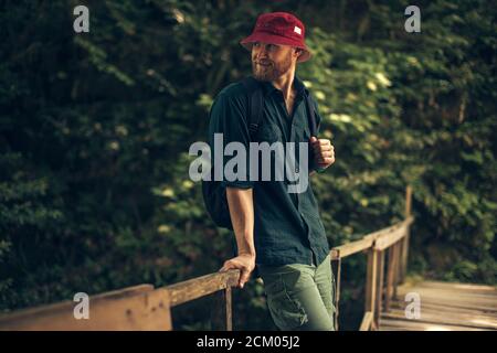 Randonneur en chapeau rouge et vêtements de voyage avec sac à dos debout sur le vieux pont en bois entouré d'une forêt tropicale luxuriante Banque D'Images
