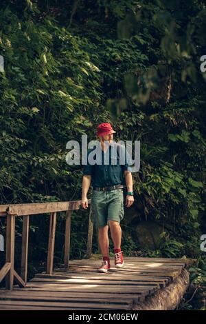 Jeune caucasien Hiker dans le chapeau rouge et les vêtements de voyage avec sac à dos sur le vieux pont en bois entouré de végétation luxuriante forêt tropicale Banque D'Images