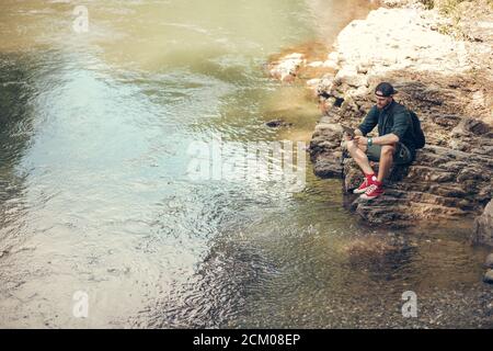 Vue de dessus d'un jeune aventurier reposant, voyageant seul dans les forêts sauvages d'été, Bearded Hiker utilisant une tablette sur la nature tout en étant assis sur le lac s pierreux Banque D'Images