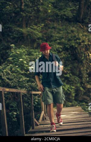 Jeune caucasien Hiker dans le chapeau rouge et les vêtements de voyage avec sac à dos sur le vieux pont en bois entouré de végétation luxuriante forêt tropicale Banque D'Images