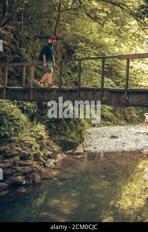 Jeune caucasien Hiker dans le chapeau rouge et les vêtements de voyage avec sac à dos sur le vieux pont en bois entouré de végétation luxuriante forêt tropicale Banque D'Images
