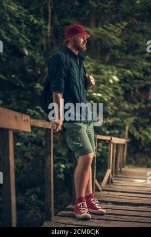 Jeune caucasien Hiker dans le chapeau rouge et les vêtements de voyage avec sac à dos sur le vieux pont en bois entouré de végétation luxuriante forêt tropicale Banque D'Images