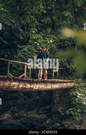 Jeune caucasien Hiker dans le chapeau rouge et les vêtements de voyage avec sac à dos sur le vieux pont en bois entouré de végétation luxuriante forêt tropicale Banque D'Images