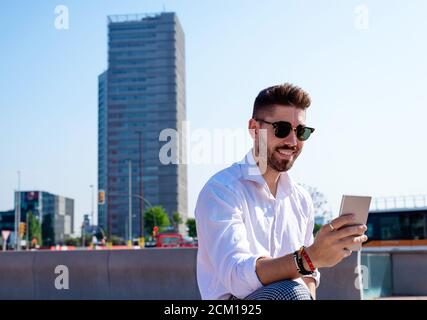 Un jeune homme barbu est assis sur un banc à l'extérieur, contre un verre moderne bâtiment Banque D'Images