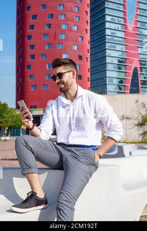 Un jeune homme barbu est assis sur un banc à l'extérieur, contre un verre moderne bâtiment Banque D'Images