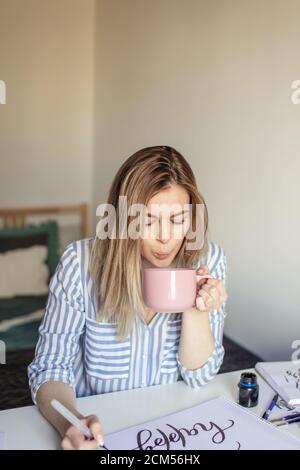 Gros plan de la femme écrivant de belles notes sur le papier sur le bureau blanc tout en prenant un café ou du thé Banque D'Images