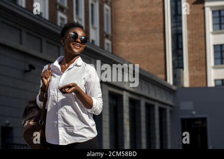 Photo d'été en plein air d'une femme d'affaires afro-américaine joyeuse vêtue d'une chemise blanche de base, tenant un sac à dos derrière son épaule, se précipitant jusqu'à la réunion Banque D'Images