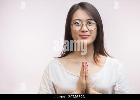 Femme asiatique à poils longs, avec lunettes optiques, mains dans le geste de Namaste exprimant un accueil chaleureux et amical, prise de vue en studio isolée sur wh Banque D'Images