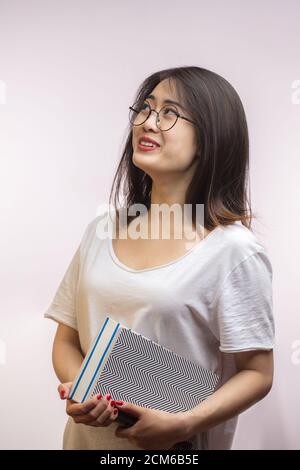 Cheerful student asiatique femme portant des lunettes, habillé en t-shirt blanc et intéressante lecture livre drôle sur fond blanc isolée à st Banque D'Images