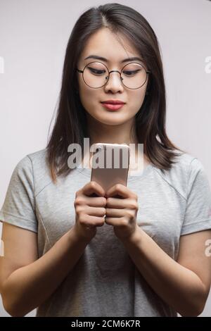 Adorable femme asiatique étudiant utilisant un smartphone pour commander de la pizza en ligne, regardant attentivement le smartphone, vêtu d'un t-shirt gris, isolé sur blanc Banque D'Images