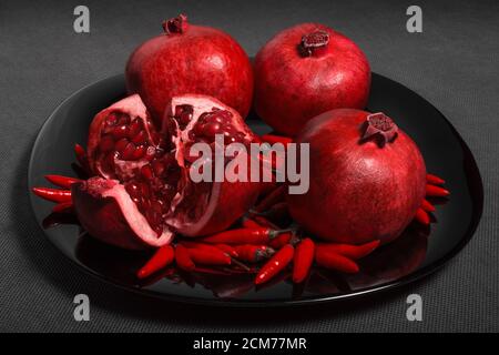 Fruit grenade et mini poivre rouge sur une assiette noire, sur un platre gris neutre, horizontal Banque D'Images