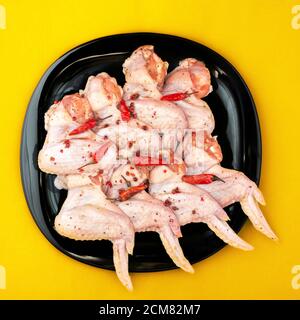 Ailes de poulet crues aux épices. Poulet cru mariné au piment fort, sur une assiette noire, fond jaune vif Banque D'Images