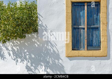 La fenêtre d'une maison grecque sur un mur blanc Avec bordure jaune et volets en bois bleu sur un Soleil jour Banque D'Images