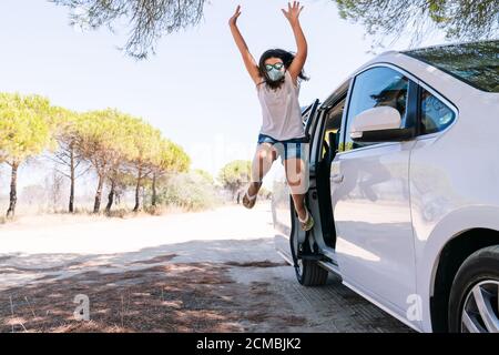 Fille saute de la porte de voiture avec un visage masque et lunettes de soleil heureux avec ses bras ouverts pour avoir nous avons pu profiter des vacances d'été après le c Banque D'Images