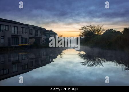 Une rangée de maisons entourant un lac brumeux rempli de réflexions des maisons et des arbres au lever du soleil, le Cap, Afrique du Sud Banque D'Images