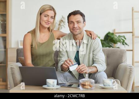 Portrait d'un jeune couple souriant à l'appareil photo tout en payant achats en ligne avec carte de crédit à l'aide d'un ordinateur portable Banque D'Images