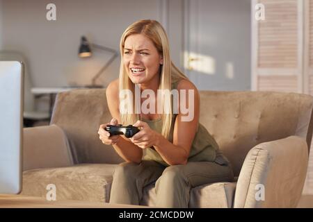 Jeune femme blonde assise sur un canapé à la maison et jouant jeu informatique avec joystick Banque D'Images