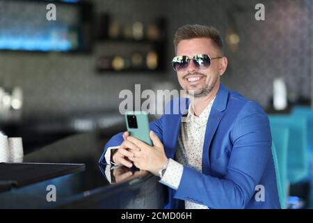 Un homme heureux en veste bleue avec barbe et lunettes de soleil s'assoit au bar et attend. Banque D'Images