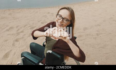 Subport pour personnes handicapées. Jeune femme en fauteuil roulant sur la côte sablonneuse de la rivière montrant le cœur avec les mains. Photo de haute qualité Banque D'Images