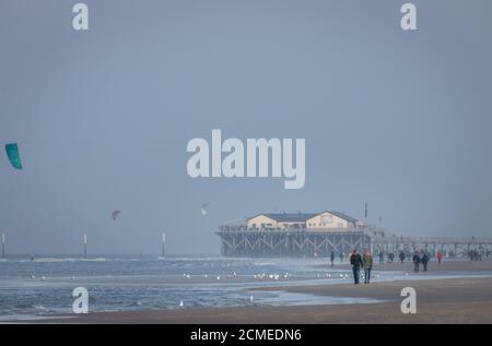 Personnes marchant le long de la plage de la mer du nord à Sankt Peter-Ording on une journée ensoleillée en automne avec des maisons de pilotis dans le arrière-plan dans le ciel Banque D'Images