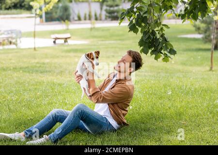 jeune homme en chemise marron et jean tenant le jack russell chien terrier assis sur la pelouse Banque D'Images