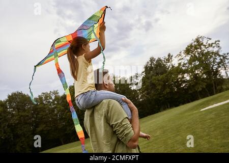 Passer du temps avec un super papa. Vue arrière d'une petite fille assise sur les épaules de son père et jouant avec un cerf-volant coloré tout en se tenant dans le parc sur un Banque D'Images