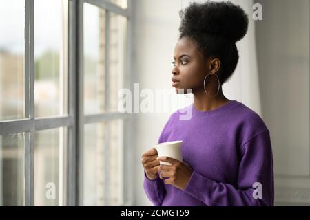 Pensive Afro-américaine jeune femme millénaire dans les bijoux à la mode grandes boucles d'oreilles porter le chandail violet pensée, regardant à travers la fenêtre, tenant blanc Banque D'Images